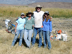 A tall man with a gray beard stands arm-in-arm with three shorter women, all dressed in jeans and T-shirts, on a dry grass plain