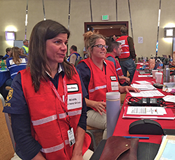two women wearing red vests, with other people in an incident command post