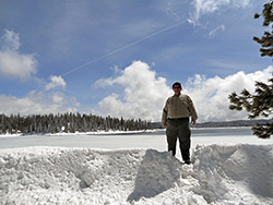 A large man stands in snow, with a lake and forested hills behind him