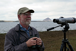 A man with a gray beard and glasses, wearing a baseball cap and dark gray jacket, stands next to a spotting scope on a tripod, near Morro Bay, with Morro Rock in the background.