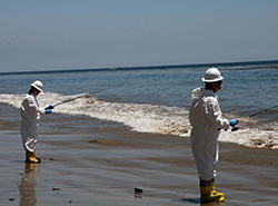 Two adults wearing hard hats and chemical-resistant jumpsuits and boots fish for samples in shallow surf during an oil spill