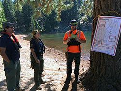 A woman in uniform and two men stand under pines near a river