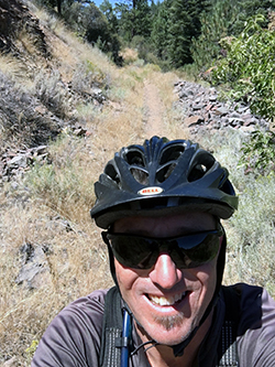Face of a smiling Caucasian man wearing a bicycle safety helmet, with a mountain bike trail and forest behind him