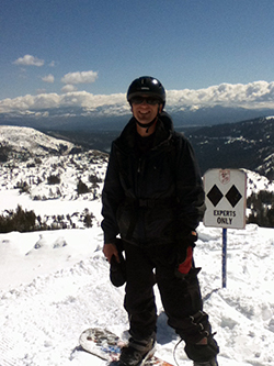 A snowboarder dressed in black stands on a peak in a snow-covered mountain range, in front of an “Experts Only” sign