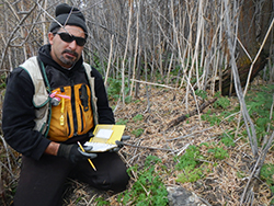 A man with a dark goatee, wearing black with an orange safety vest, kneels among dead reeds and low vegetation, holding a field notebook