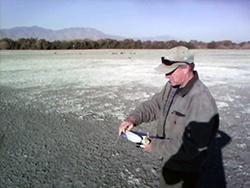 A man with an electronic device in hand stands in a dry river bed with dried salt residue in background