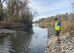Scientist, Doug Killam standing on the rocky banks for the Sacramento River with trees and shrubs in background