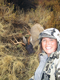 an immobiilized buck with an eye cover lies behind a pretty young woman wearing camouflage