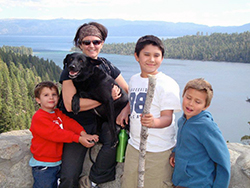 a woman holding a black labrador retriever poses with three young boys, with Emerald Bay behind them