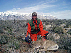 Man in orange jumpsuit kneels in sagebrush with a deer that's hobbled and blindfolded 