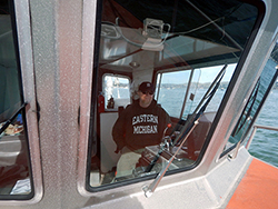 a man wearing a navy blue sweatshirt and baseball cap at the helm of a small research vessel