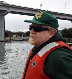 a middle-aged man wearing an orange life-vest and green baseball cap, with bay water and a concrete bridge in background