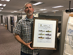 Man wearing gray plaid shirt holding frame with certificate depicting trout standing in office.