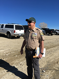 Man wearing CDFW uniform, sunglasses, and green ball cap standing in dirt parking lot holding clipboard.