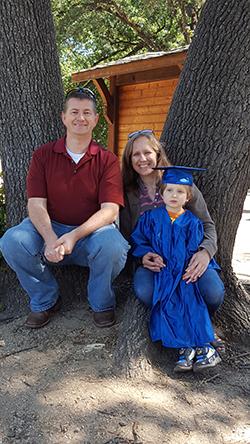 Smiling man wearing red shirt and blue jeans with smiling woman with arms around young child wearing blue graduation cap and gown in front of tree.