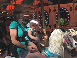 Smiling woman wearing wide brim hat, sunglasses, shorts and blue tshirt on merry go round horse holding baby in front facing baby carrier