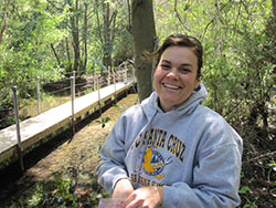 Smiling woman wearing grey sweatshirt outside in forested area with footbridge in background