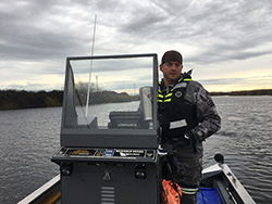 Man wearing gray camo foul weather gear, backward ball cap, and black life vest with yellow reflectors behind helm on fishing boat on water. Cloudy skies and grass-covered levy in background