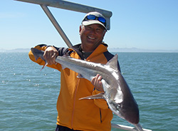 Smiling man on boat wearing yellow jacket and ballcap with sunglasses holding up shark in both hands. Water in background.