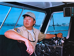 Man wearing beige official Department of Fish and Wildlife uniform with beige ball cap inside boat at helm.