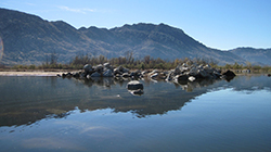 Calm lake facing a pile of large rocks partially submerged with mountains in the background