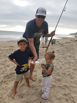 White male wearing blue t-shirt, ball cap, and sunglasses holding fishing rod in one hand and small fish in another standing on beach next to young girl and young boy. Overcast sky and water in background.