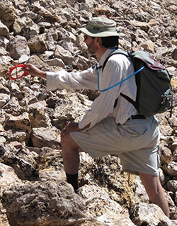Man wearing beige fishing hat, khaki pants, white long sleeved shirt, and backpack on rocky slope holding round red item