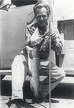 Black and white image of a man crouched in front of truck, holding a large fish in one hand and measuring stick in other hand. Man is wearing plaid collared shirt and pants.