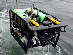Black and green machine with two yellow tanks on top above water, suspended by chain.