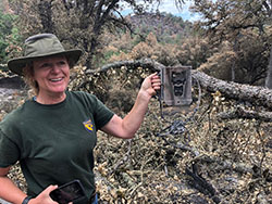 Woman in green shirt and green fishing hat holding up burned electronic box with fallen tree and trees in background.