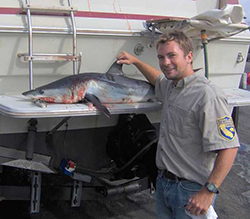 Smiling man wearing khaki shirt with CDFW arm patch with hand on dorsal fin of small shark on stern of boat.