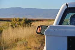 partial view of white pickup truck with face in sideview mirror. Grassland and mountain in background