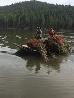 Two men wearing life jackets aboard small boat on body of water with pile of dead christmas trees. Forest and mountain in background